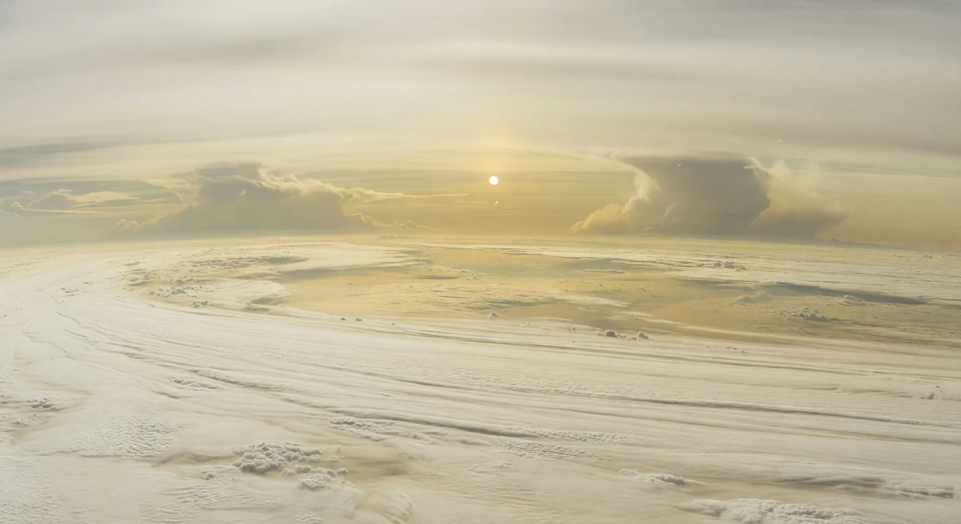 Vor Ihnen breitet sich kein Boden aus, sondern eine gewaltige, sanft gewellte Ebene aus Ammoniak-Eiswolken in Creme, Elfenbein und blassem Gold, durchzogen von flachen Mulden, niedrigen Rücken und feinen, vom Wind gestreckten Mustern, die sich bis zu einem stark gekrümmten, butterscotchfarbenen Horizont ziehen. In dieser Höhe von etwa ,5 bis 2 Bar schwebt man über den oberen Wolkendecks eines Gasriesen ohne jede feste Oberfläche; gelegentliche dunklere Öffnungen in Olive-Tan und Graubraun lassen nur ahnen, dass weit darunter tiefere Wolkenschichten aus Ammoniumhydrosulfid liegen. Das Licht ist schwach und kalt, denn die ferne Sonne erscheint hier nur als kleines, scharfes weiß-goldenes Scheibchen hinter dichtem photochemischem Dunst, dessen Aerosole den Himmel in matte Gelb-, Beige- und Grauweißtöne auflösen. Trotz der stillen Weite verraten riesige, weich aufragende Wolkenwände in der Ferne die planetare Dimension dieser Atmosphäre, in der alles Landschaftliche aus Schichtung, Konvektion und Strömung besteht — eine ruhige, fremdartige Welt aus Wolken statt Gestein.