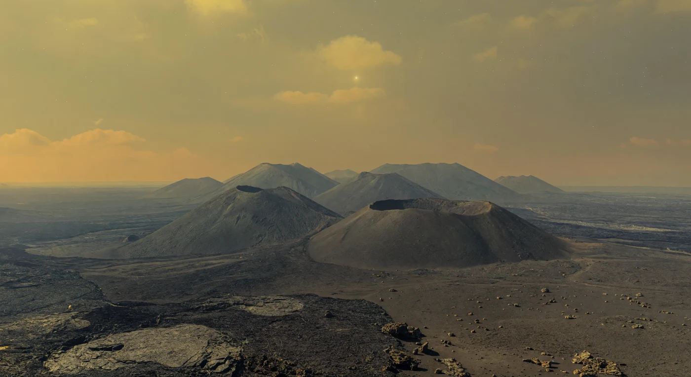 Davanti a te si estende una pianura di lave basaltiche scure, spezzata da lastre fratturate, blocchi angolosi e superfici che alternano texture lisce simili al pāhoehoe e colate più ruvide, mentre all’orizzonte emergono più vulcani a scudo adiacenti, larghi e poco inclinati, fusi in un unico massiccio dalle proporzioni immense. Sulle loro sommità si distinguono depressioni calderiche e bordi collassati, appena visibili attraverso una foschia densa color giallo-ocra che appiattisce il paesaggio con una luce diffusa e quasi priva di ombre. Queste forme raccontano una superficie costruita da enormi eruzioni effusive di lave basaltiche, poi deformata da fronti di colata, basse scarpate tettoniche e rughe del terreno, in un ambiente dove l’atmosfera di anidride carbonica, opprimente per pressione e temperatura, altera l’aspetto delle rocce e cancella ogni contorno netto. Il cielo, uniforme e senza dettagli, incombe come un soffitto di nubi e gas, così che stare qui significa trovarsi in un mondo vulcanico soffocante e smisurato, dove ogni rilievo appare vicino ma in realtà si perde in una distanza resa ambigua dalla foschia.