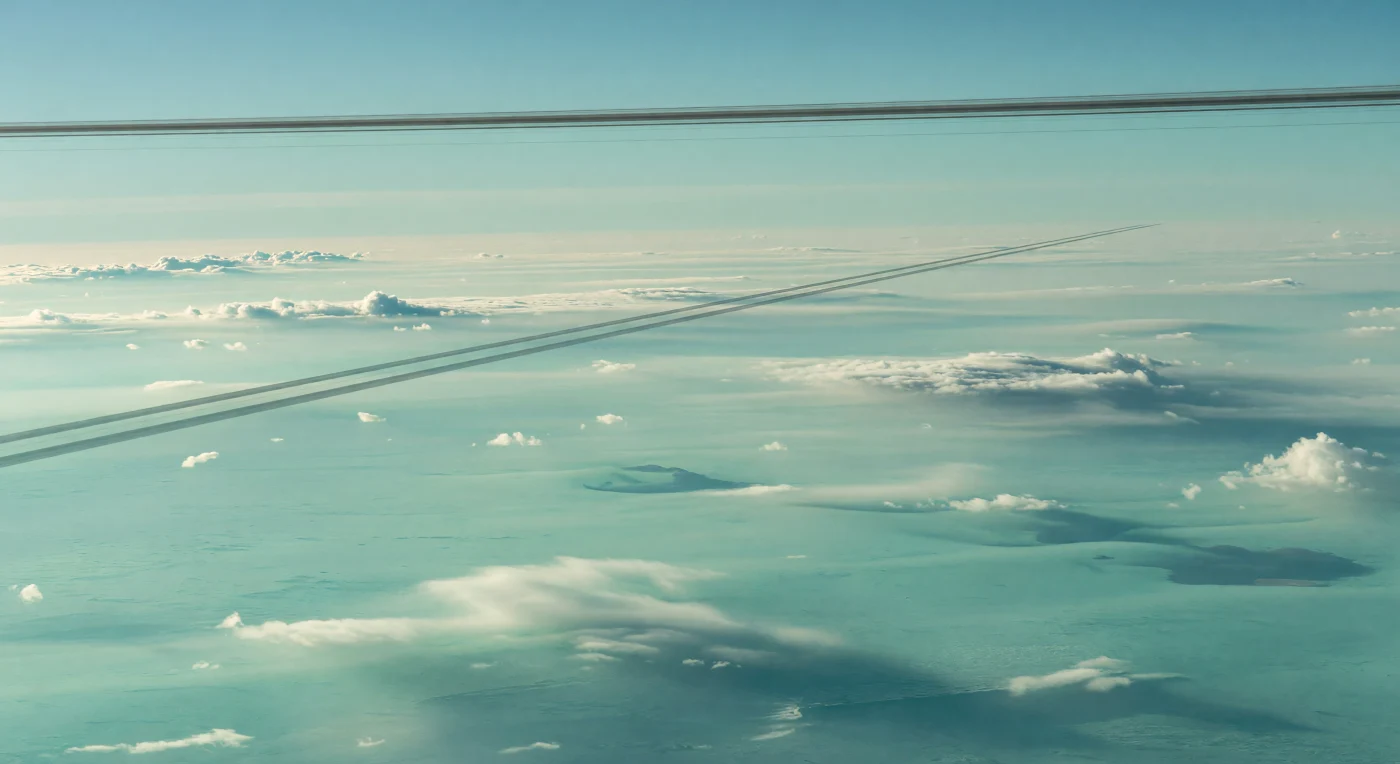 Suspenso no alto da troposfera superior, o olhar encontra não um solo, mas uma planície atmosférica imensa de nuvens verde-água e névoas azuladas, ondulando suavemente até se perderem na curvatura do horizonte. Em contraste impressionante com essas formas macias, finíssimas faixas cinzentas, perfeitamente retas e paralelas, cortam a camada de nuvens: são as sombras projetadas pelos anéis estreitos e escuros no equinócio, quando a geometria entre o Sol, os anéis e a atmosfera as torna visíveis como linhas de régua sobre o nevoeiro. Acima, os próprios anéis surgem como arcos delicados de carvão no céu pastel, quase apagados pela bruma rica em metano e aerossóis de hidrocarbonetos, sob uma luz solar minúscula, fria e difusa. Sem montanhas, mares ou rocha alguma, toda a paisagem é feita de gás, condensados e névoa estratificada — um cenário silencioso e vastíssimo onde a física orbital escreve linhas nítidas sobre um oceano de nuvens.