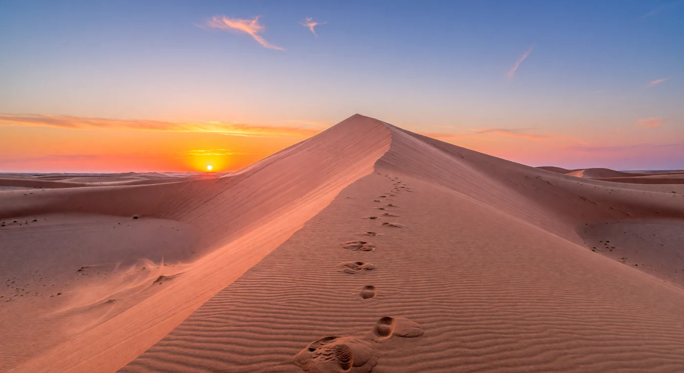From the lower flank of a towering dune, you look up a 200-meter wall of quartz-rich sand whose knife-edge crest cuts cleanly across a glowing evening sky, while tight wind ripples, faint miniature avalanche streaks, and scattered footprints reveal the constant work of desert winds on loose, well-sorted grains. Low-angle sunlight transforms the slope into bands of pink, apricot, and fiery orange that fade into a deep red-purple shadow at the base, an effect created by iron-oxide staining in the sand and warm reflected light within the dense lower atmosphere. On the horizon, the Sun hangs as a brilliant golden disk, slightly flattened by atmospheric refraction, while dust and haze near the ground intensify the vivid orange, magenta, and lavender twilight before it deepens to blue overhead. Beyond the foreground, subdued dune fields roll away into the distance with no water or vegetation to interrupt the mineral expanse, making the scene feel immense, silent, and sculpted almost entirely by air, light, and time.